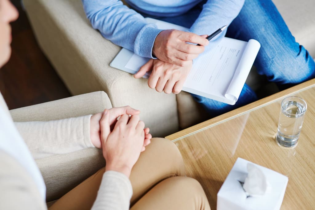 Two people sit across from each other; one holds a clipboard and pen, appearing to take notes—perhaps a private practice consultant meeting with a client. A tissue box and glass of water are on the table.